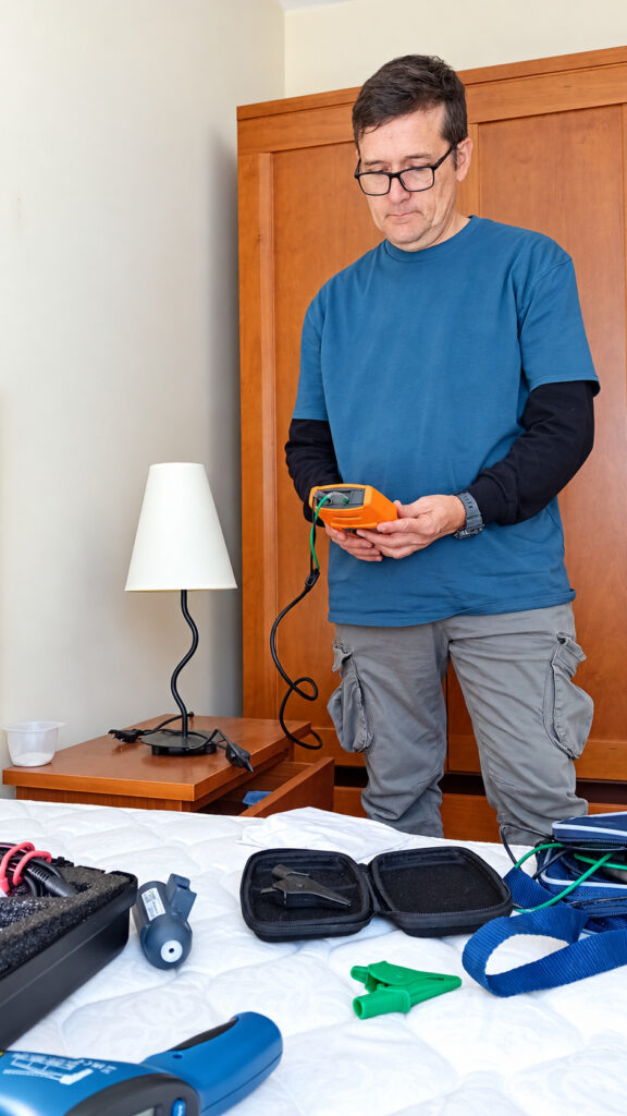 Técnico de Cemprotecta con un medidor en las manos durante una medición de resistencia de tierra en un dormitorio; el resto del equipo está sobre la cama.